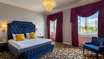 A bedroom with a large bed, a blue upholstered bench, a blue armchair, and a view of a river with ships.