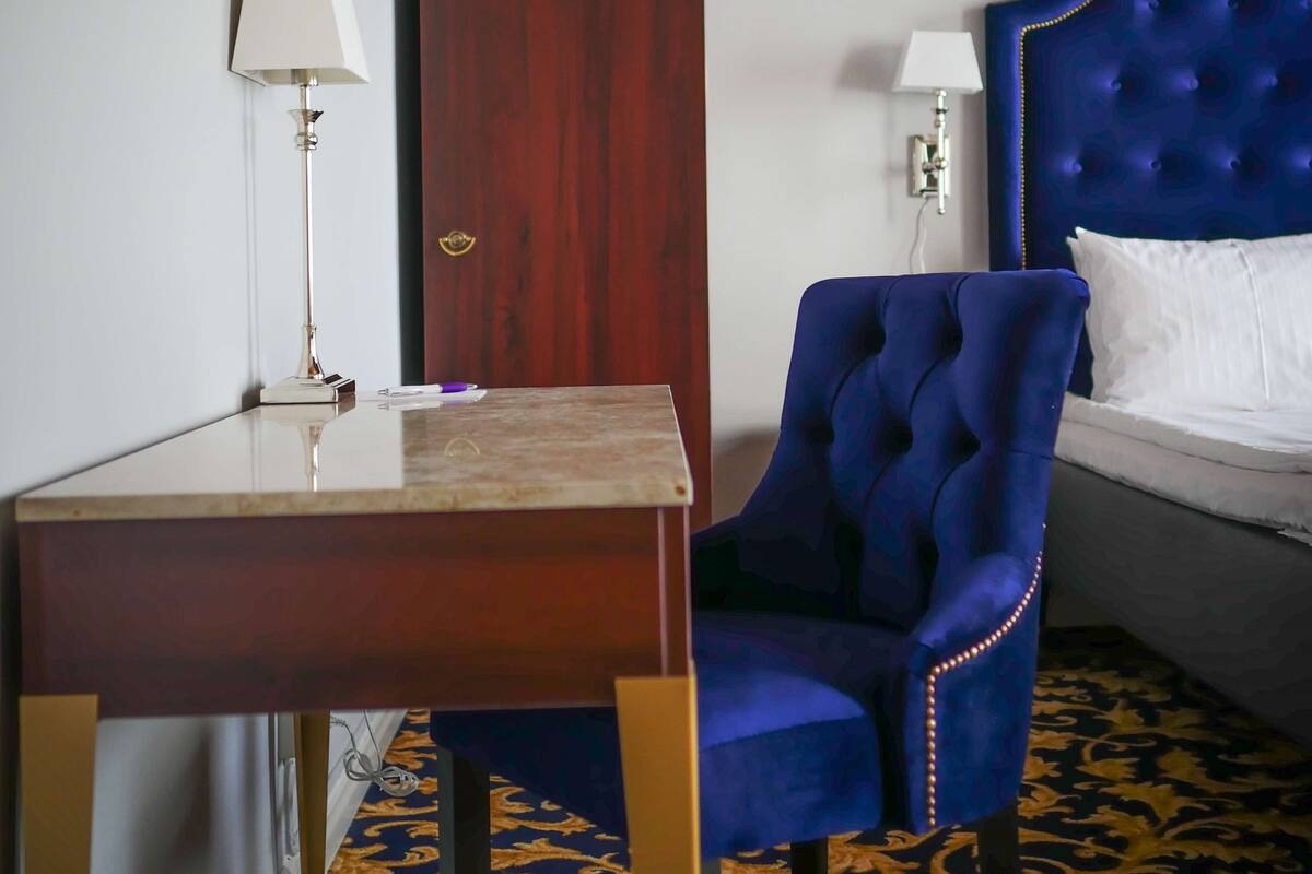 A hotel room with a wooden desk, a blue upholstered chair, a bed with a tufted headboard, and a patterned carpet.