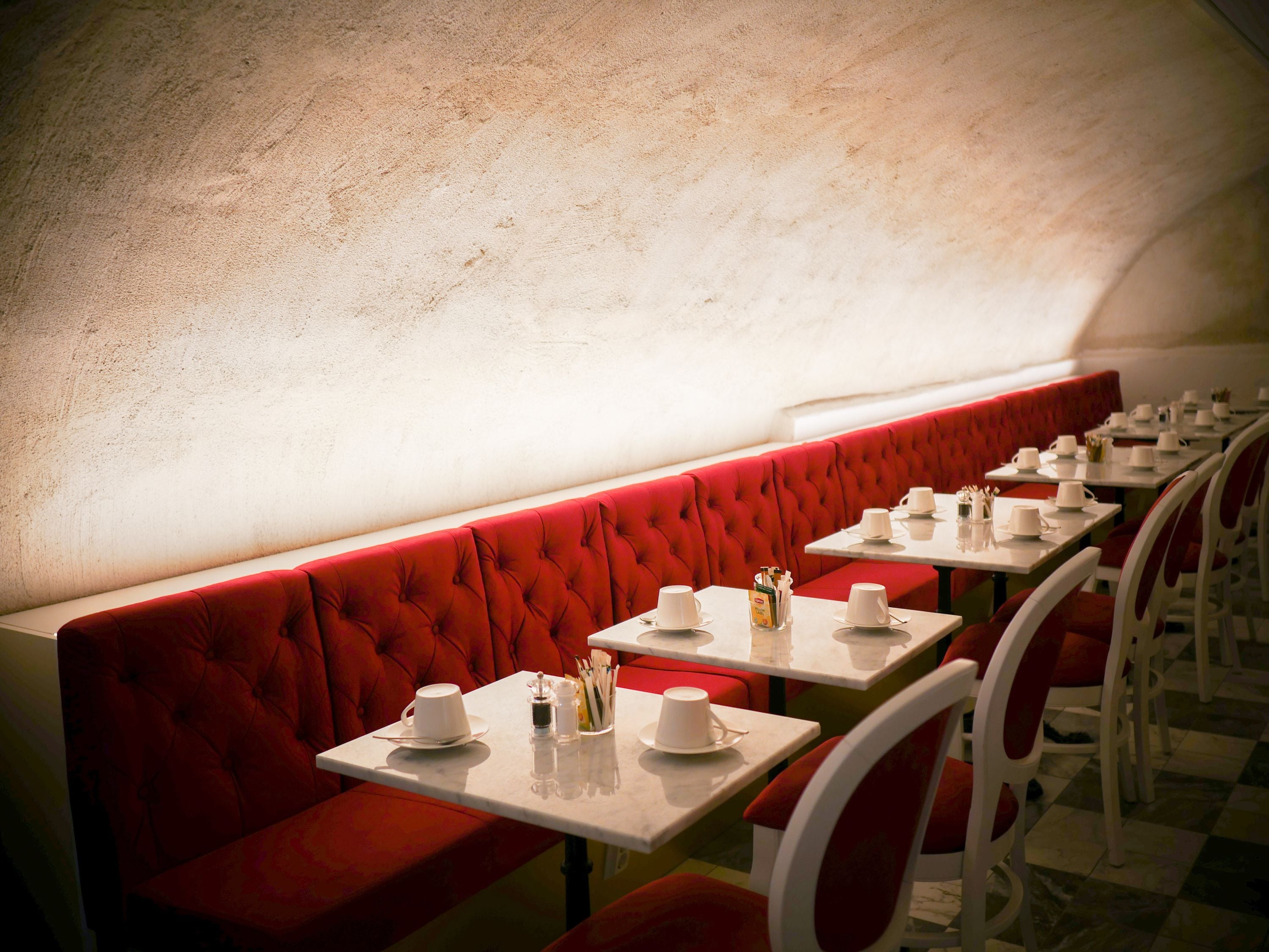 Un restaurante con asientos tapizados en rojo y mesas blancas, cada una con tazas y platillos blancos.