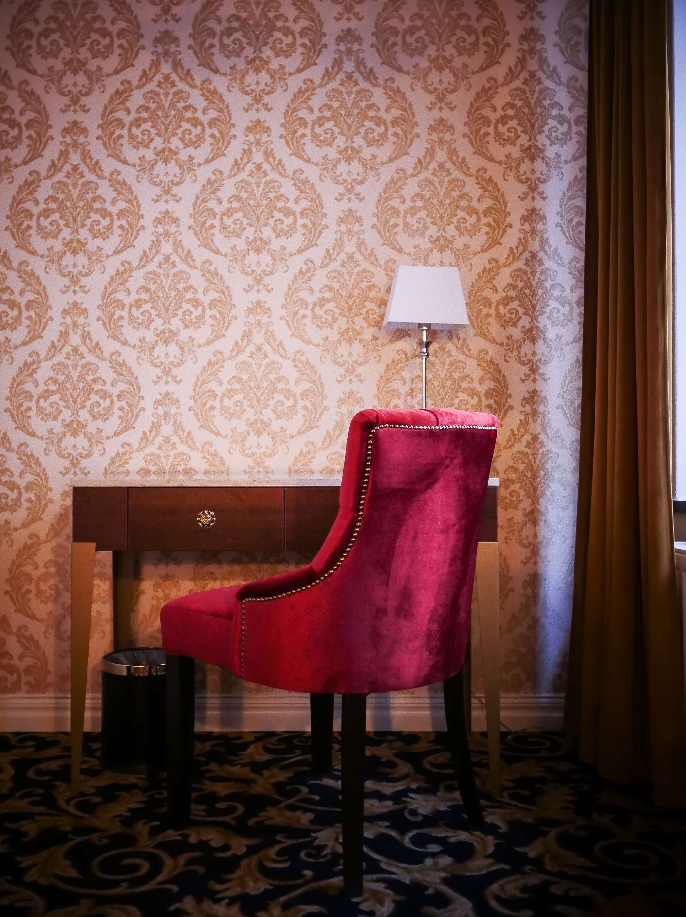 Una habitación con papel tapiz estampado, un escritorio con lámpara, una butaca de terciopelo rojo y un cubo de basura.