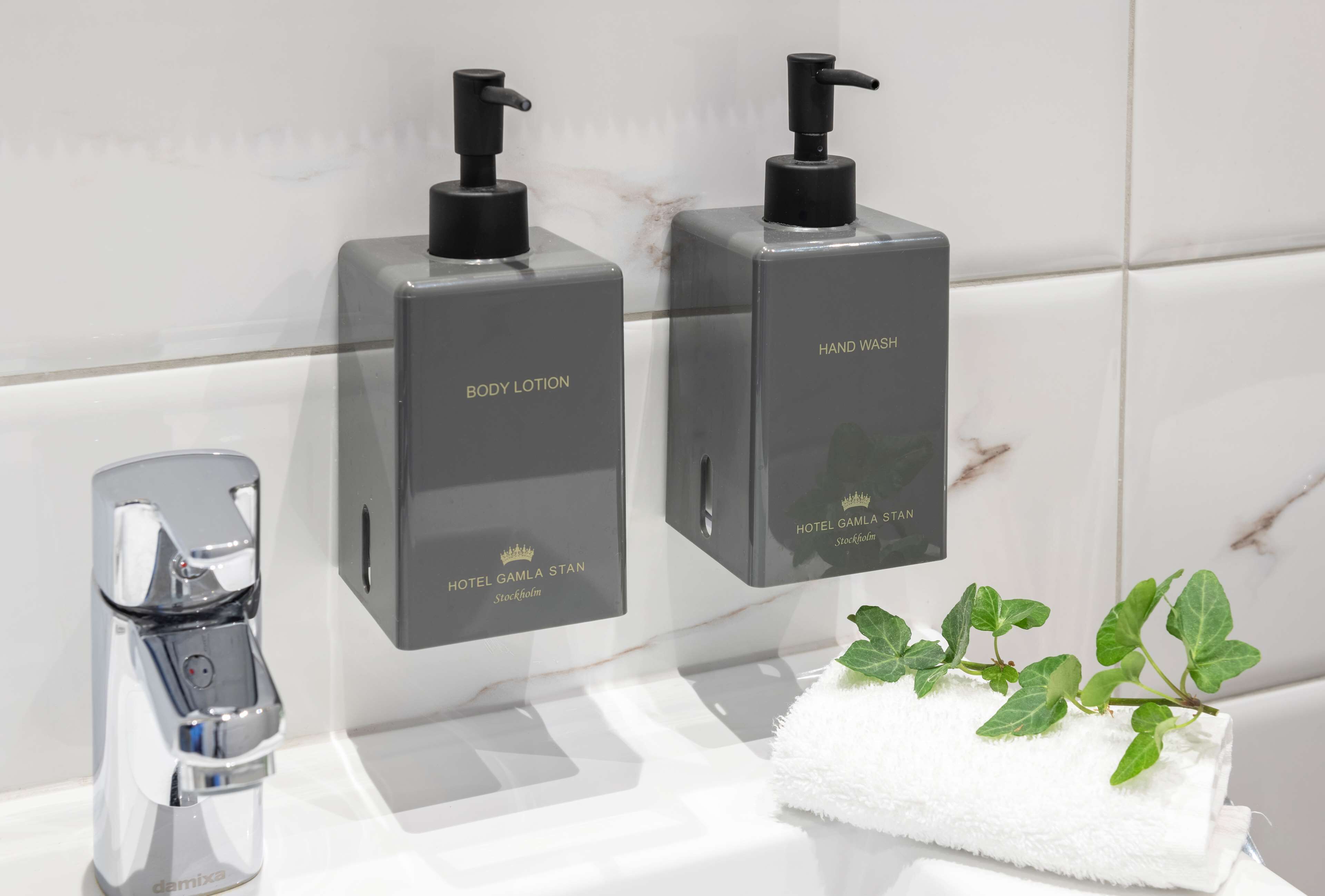 A bathroom with a modern faucet, two dispensers labeled 'BODY LOTION' and 'HAND WASH', and a towel with green leaves.
