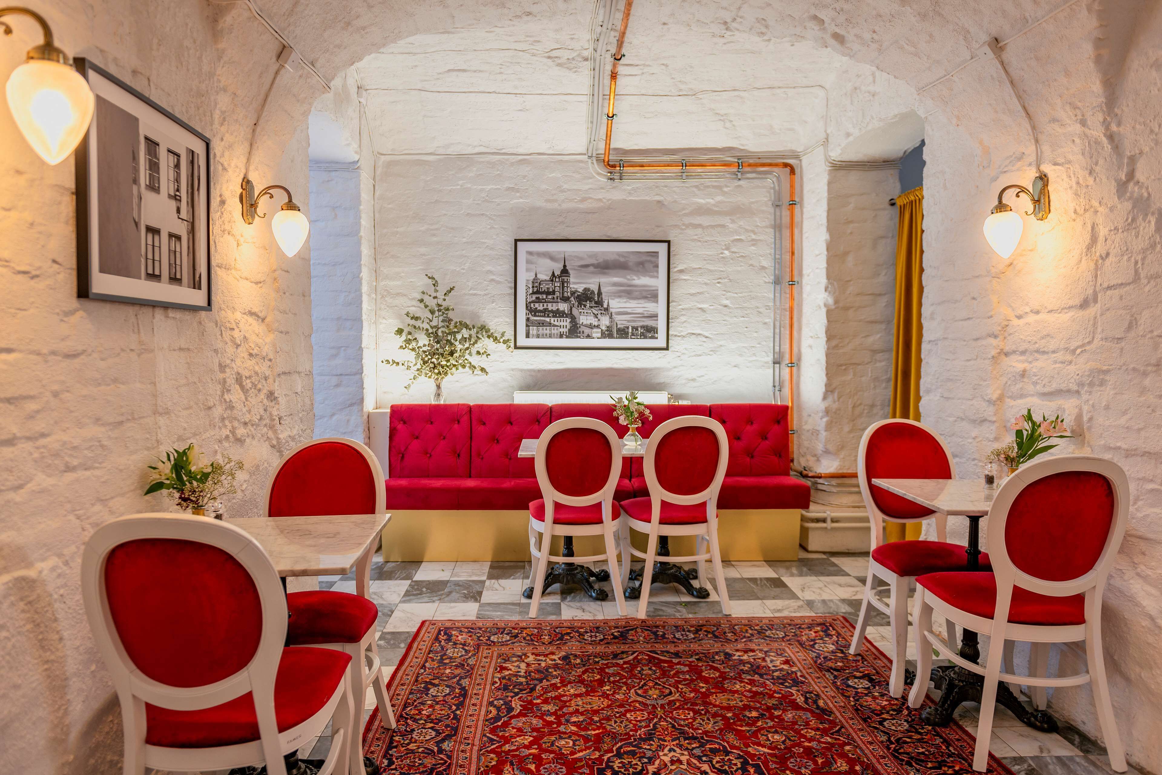 Un área de comedor con un sofá rojo, sillas blancas, un tapete estampado y cuadros en las paredes.