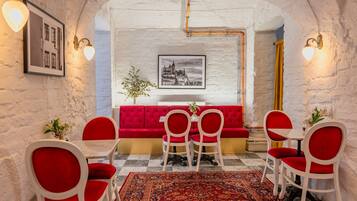 Un área de comedor con un sofá rojo, sillas blancas, un tapete estampado y cuadros en las paredes.