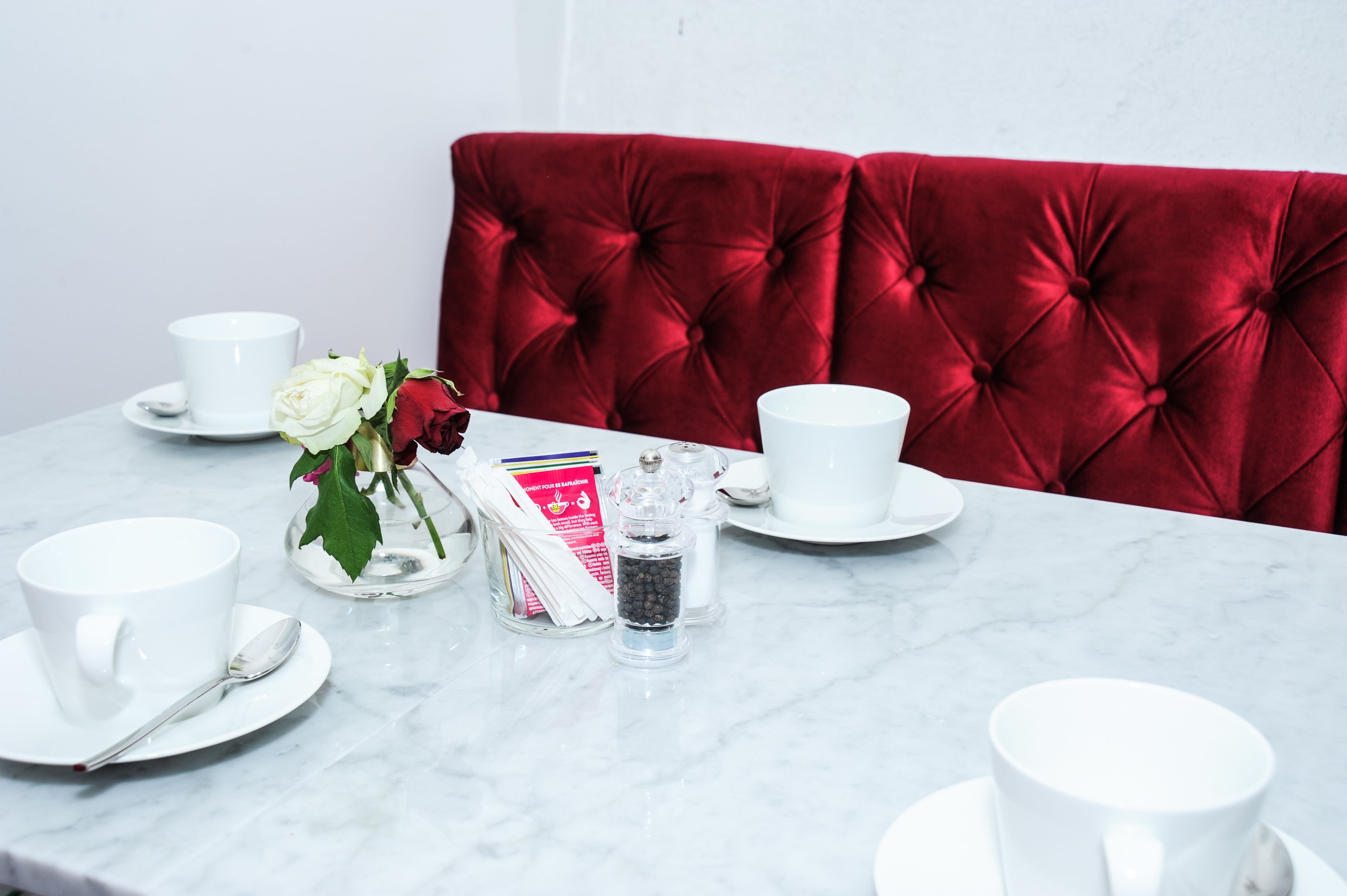 Una mesa dispuesta con tazas y platitos blancos, y una cuchara, junto con un jarrón de rosas blancas y rojas, un salero y pimentero, y un pequeño recipiente de vidrio con cuentas negras.