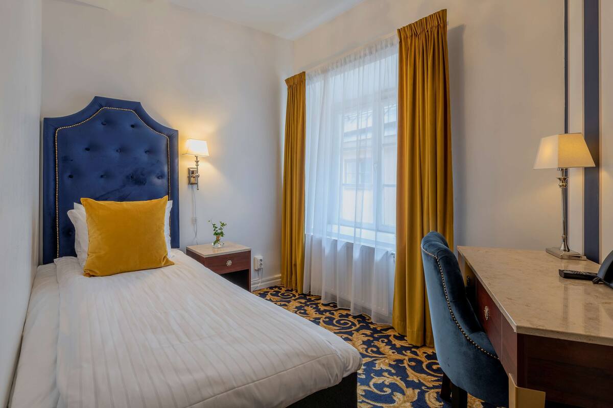 A hotel room with a large bed, a desk, and a chair. There is a window with curtains and a lamp on the desk.