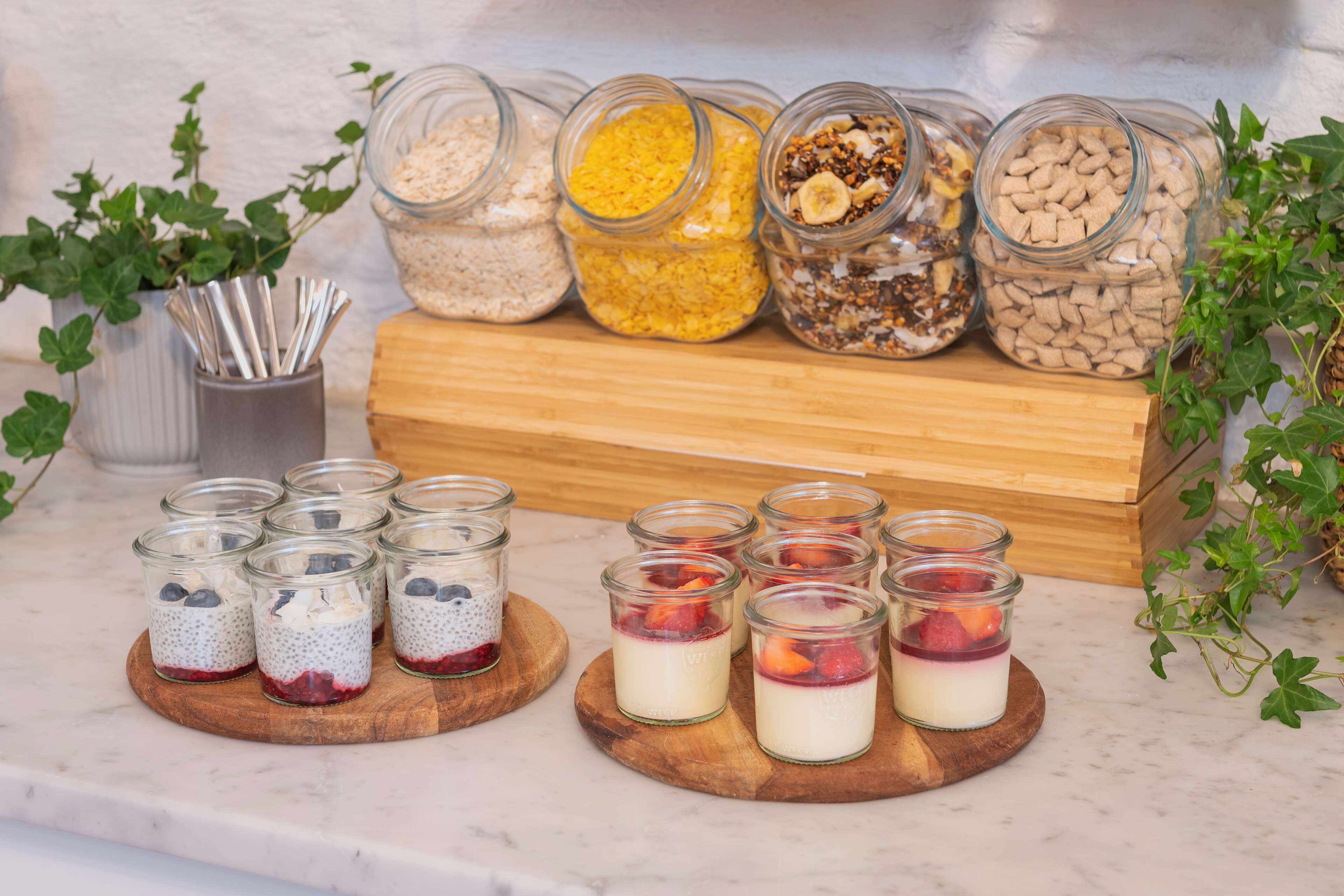 Diversos alimentos en frascos y recipientes pequeños sobre una tabla de madera, con una planta al fondo.
