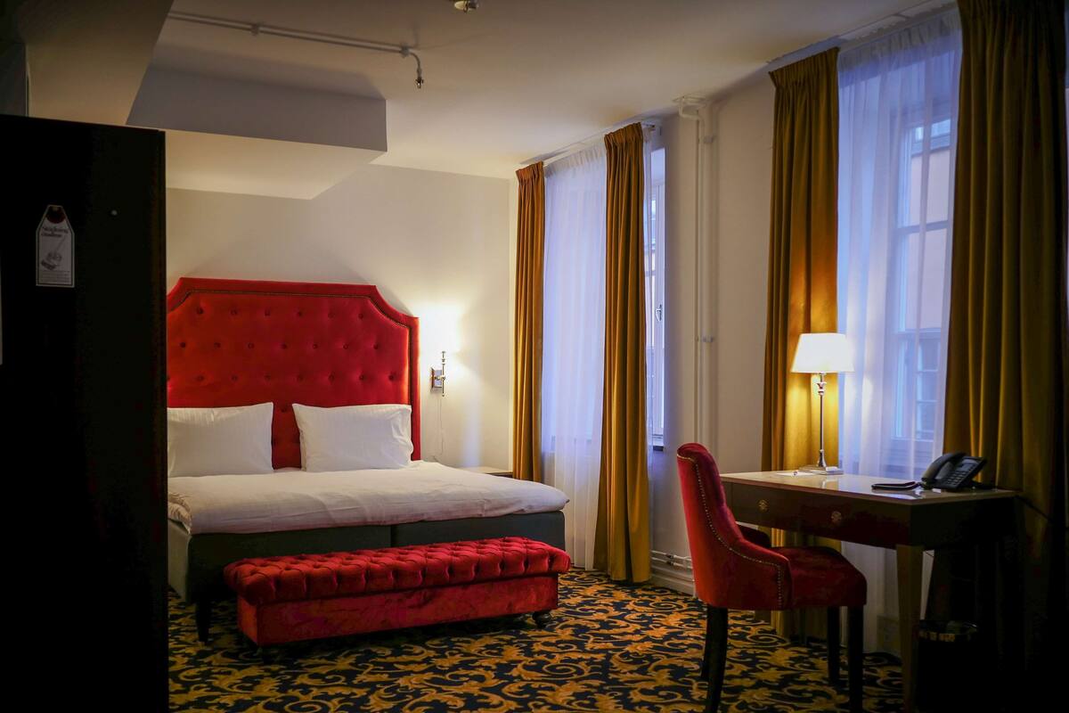 A hotel room with a red upholstered bed, a matching ottoman, a desk with a chair, and a large window with curtains.