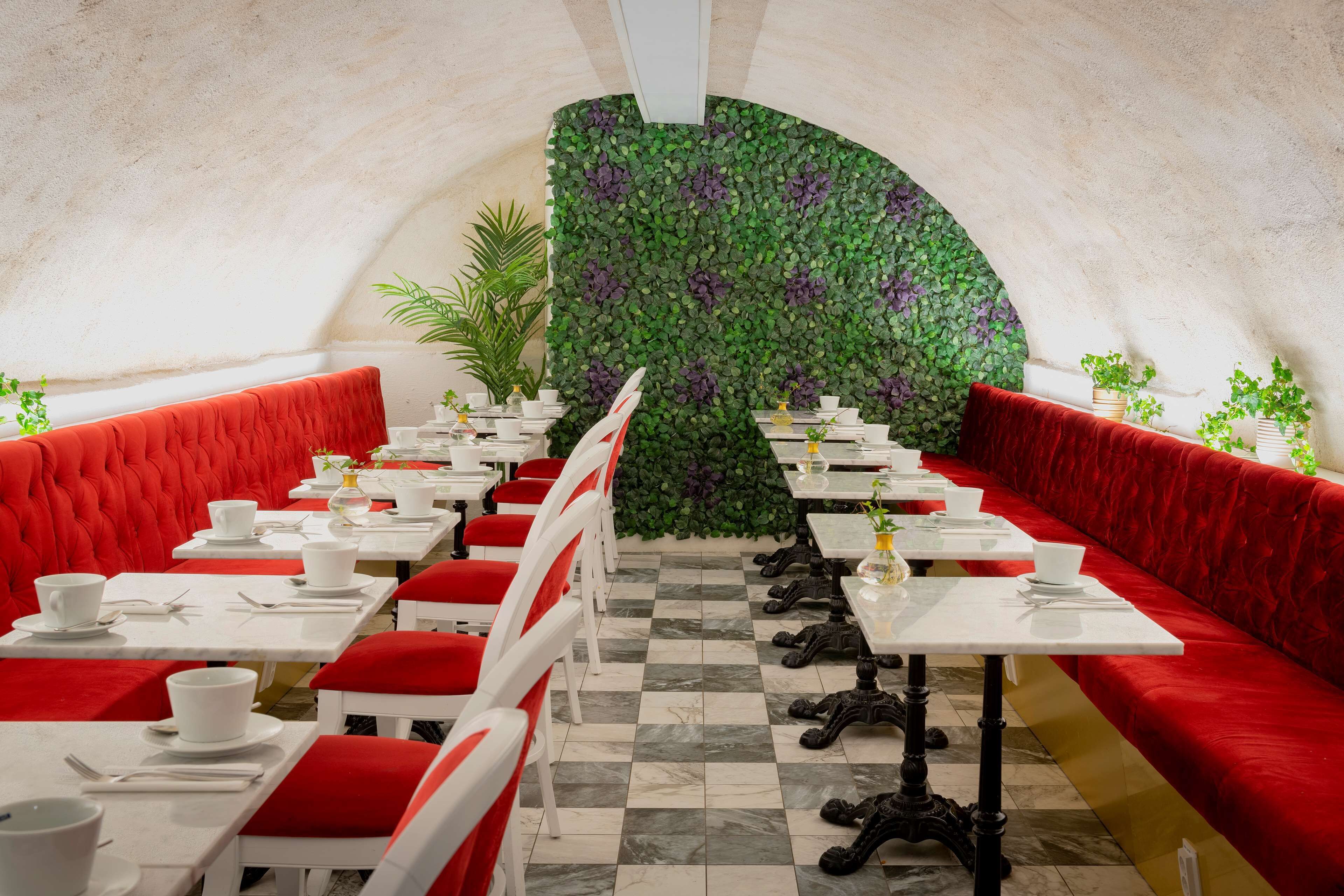 An indoor dining area with red upholstered booths, white tables, and a green leafy wall.