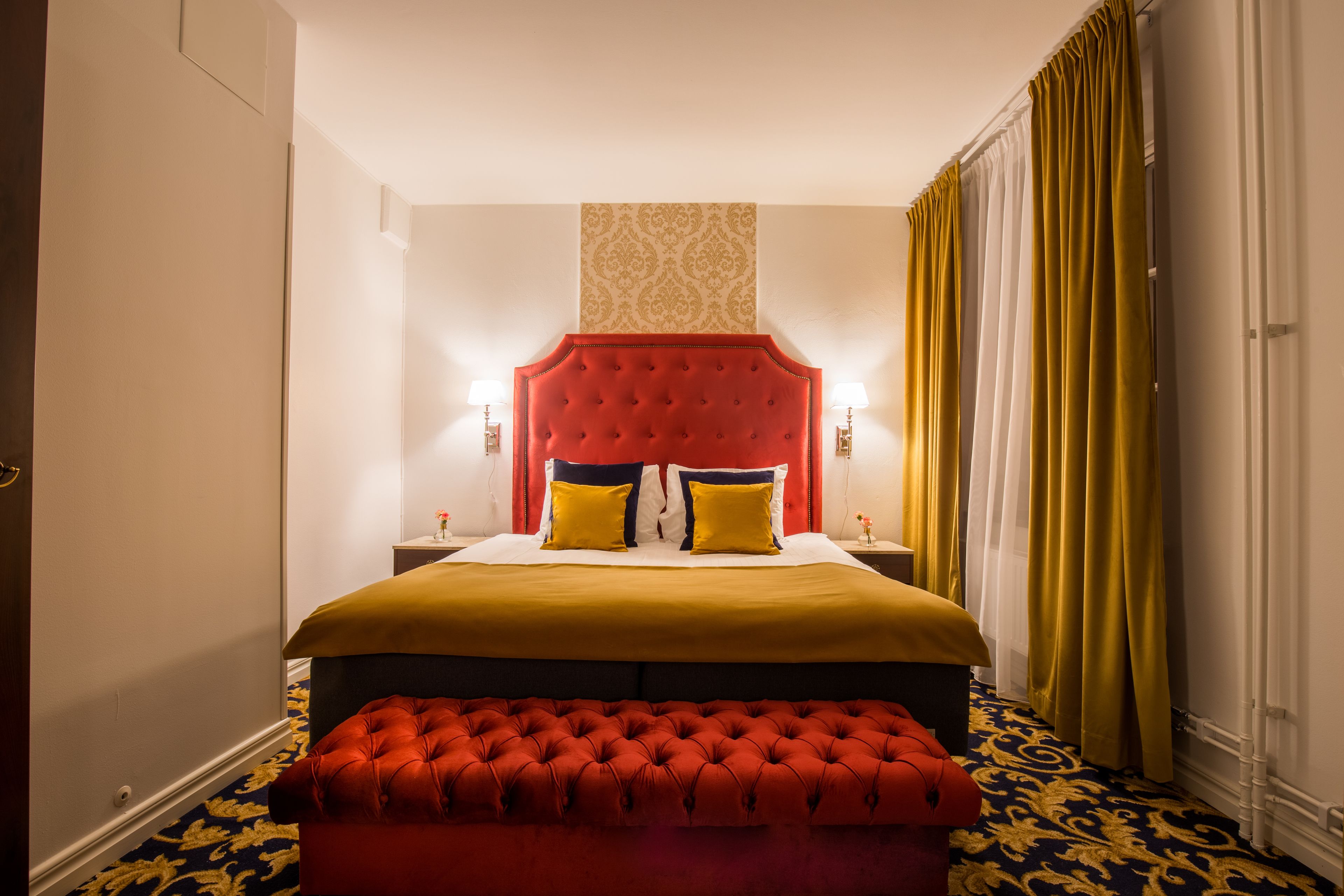A hotel room with a large bed, a red upholstered bench, and a patterned carpet.