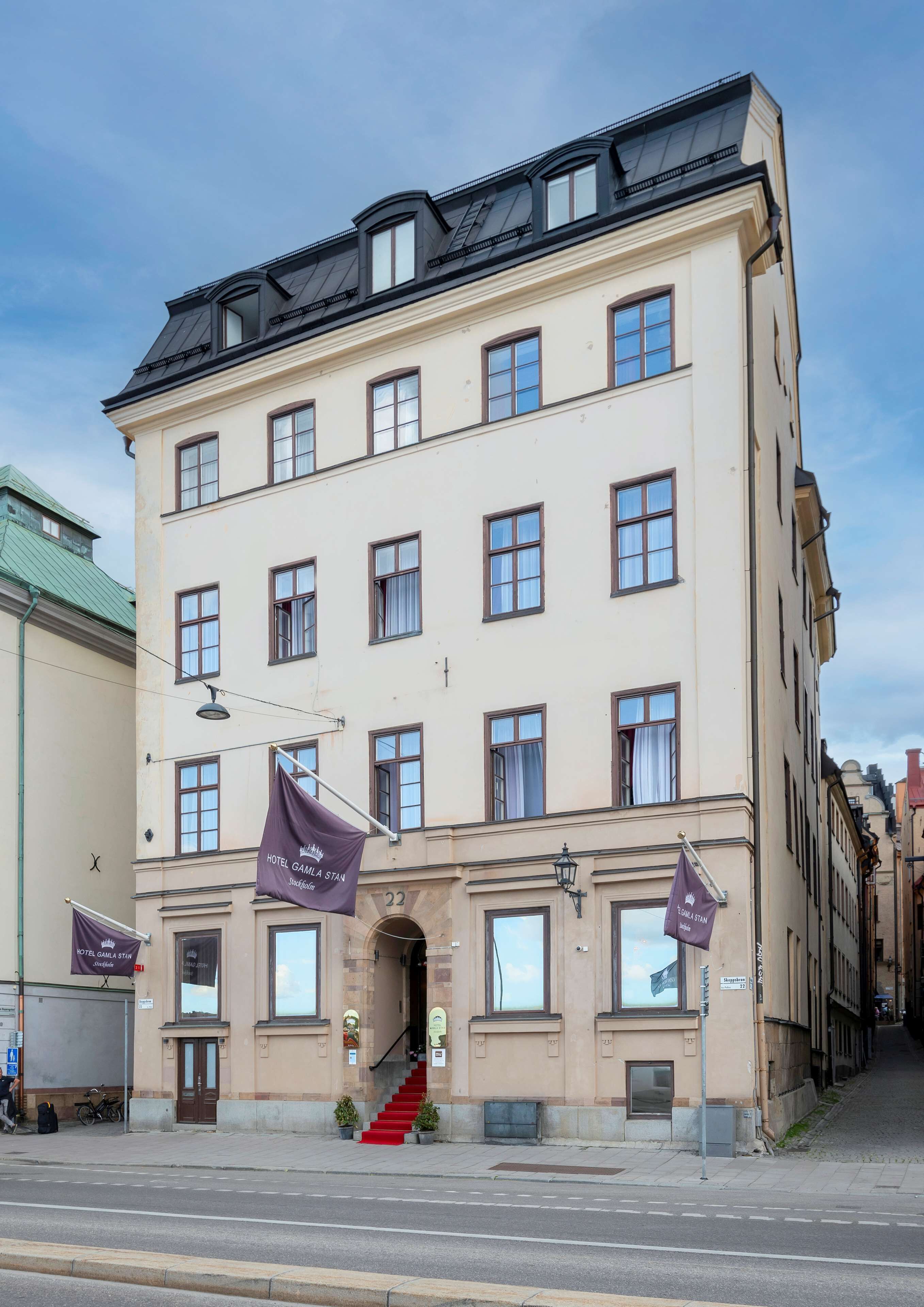 Un hotel de varios pisos con un estilo arquitectónico clásico, que cuenta con una entrada central con alfombra roja, múltiples ventanas y una bandera que indica que es un hotel.
