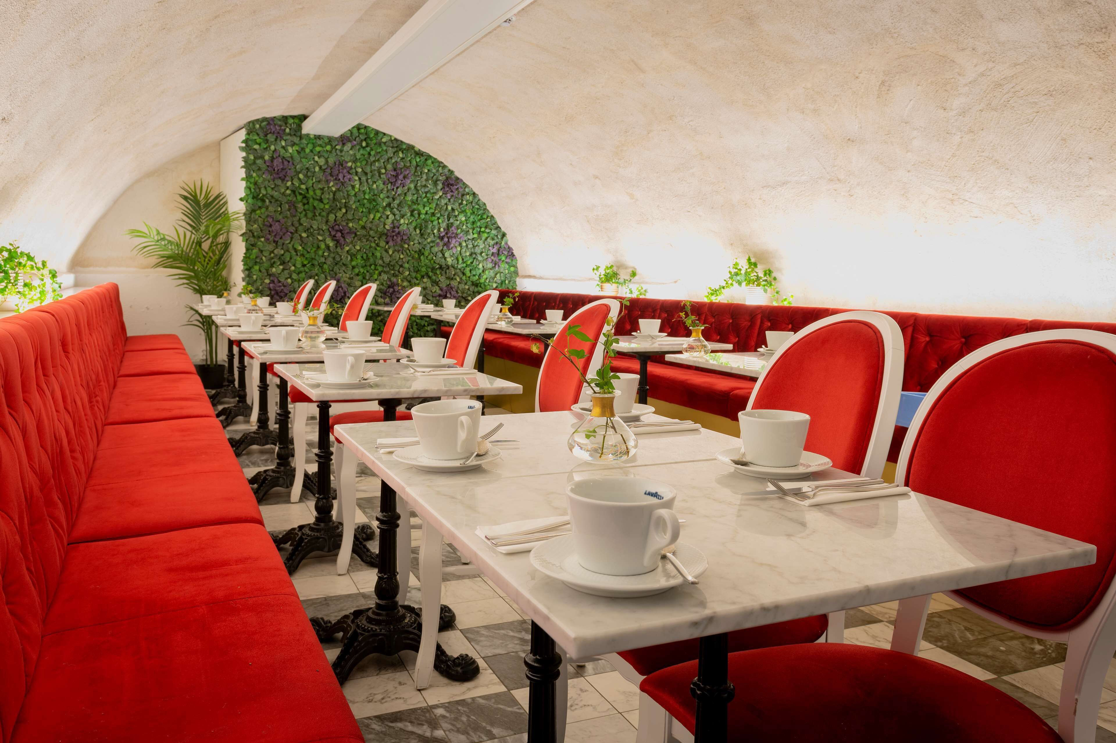Un restaurante con asientos tapizados en rojo y mesas de mármol blanco, con una pared cubierta de follaje verde y plantas en macetas.