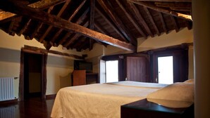 A bedroom with a large bed, wooden beams, a radiator, and a television. - Hospedería Porta Coeli (Siguenza)