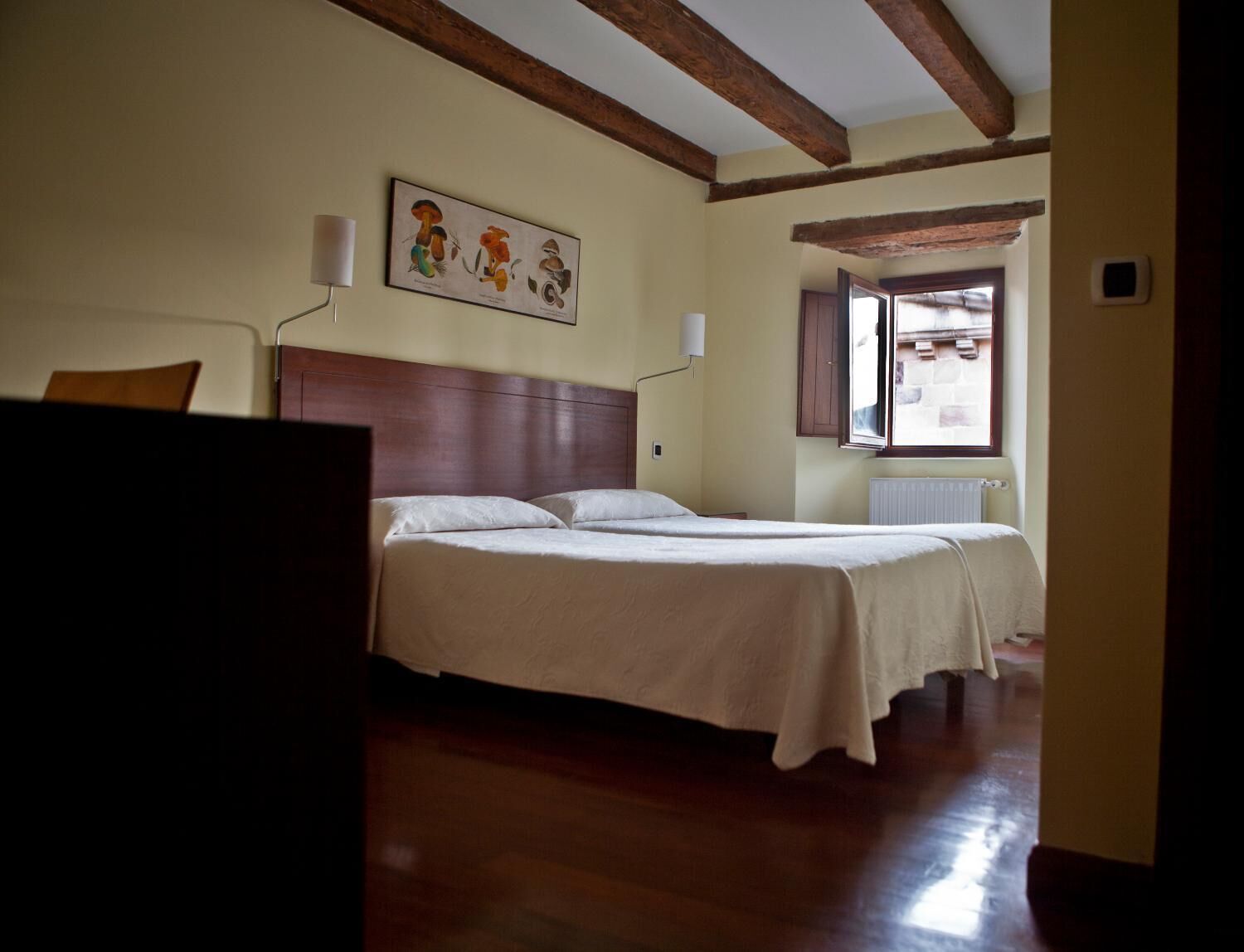 Ein Schlafzimmer mit einem Holzkopfteil, zwei Betten, einem Fenster mit Fensterläden und einem an der Wand hängenden Bild von Pilzen.