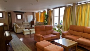 A spacious living room with a sofa set, ottomans, a coffee table, and a large window with curtains. - Hospedería Porta Coeli (Siguenza)