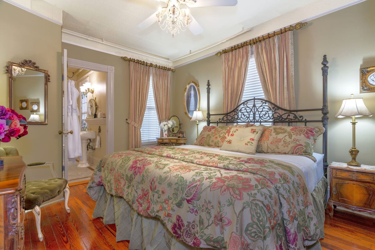 A bedroom with a large bed, a vanity with a mirror, a chandelier, and a wooden floor.