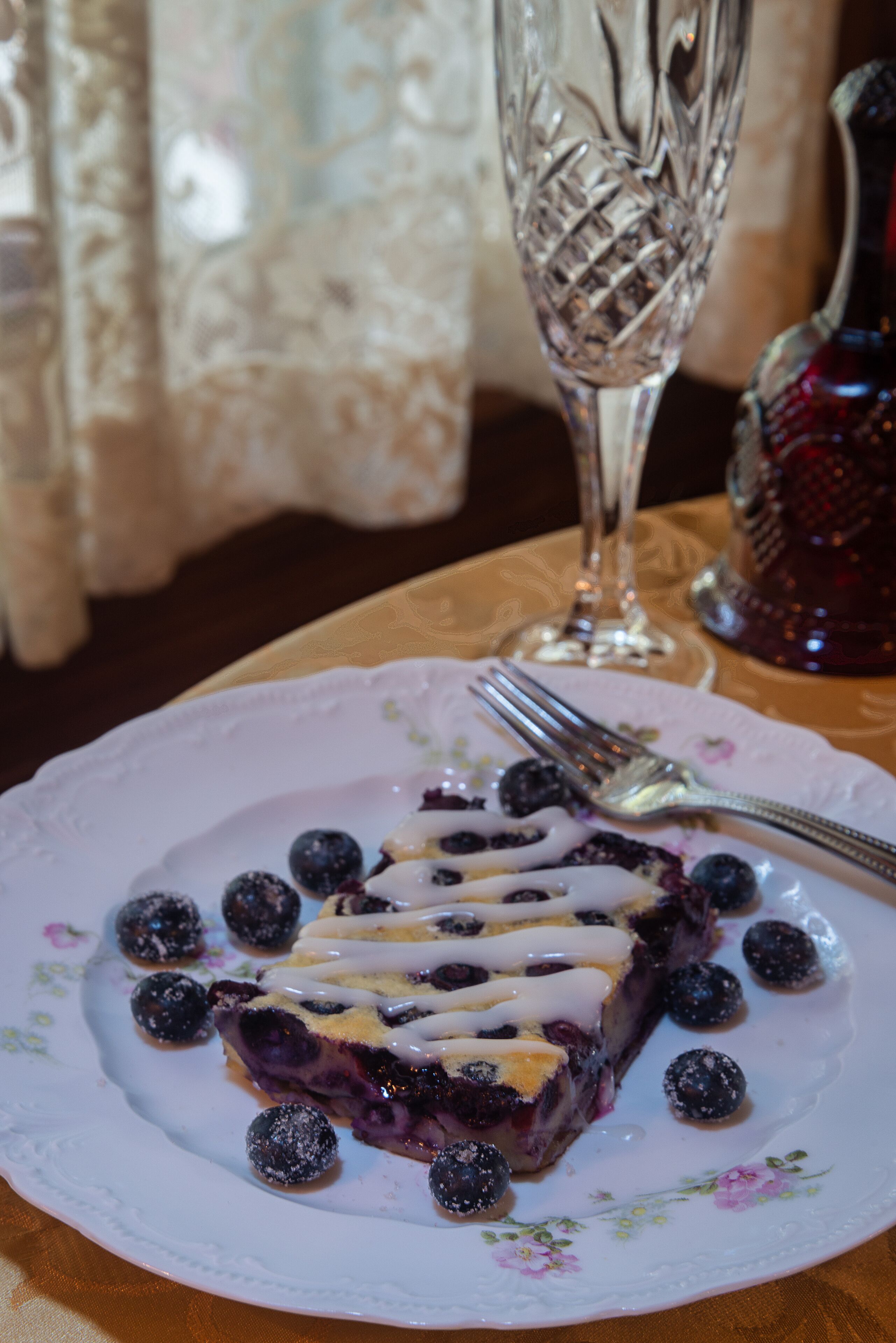Una porción de tarta azul de moras sobre un plato con motivos florales, junto con moras, un tenedor, un vaso de agua y un jarrón decorativo al fondo.