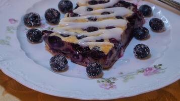 Una porción de tarta azul de moras sobre un plato con motivos florales, junto con moras, un tenedor, un vaso de agua y un jarrón decorativo al fondo.