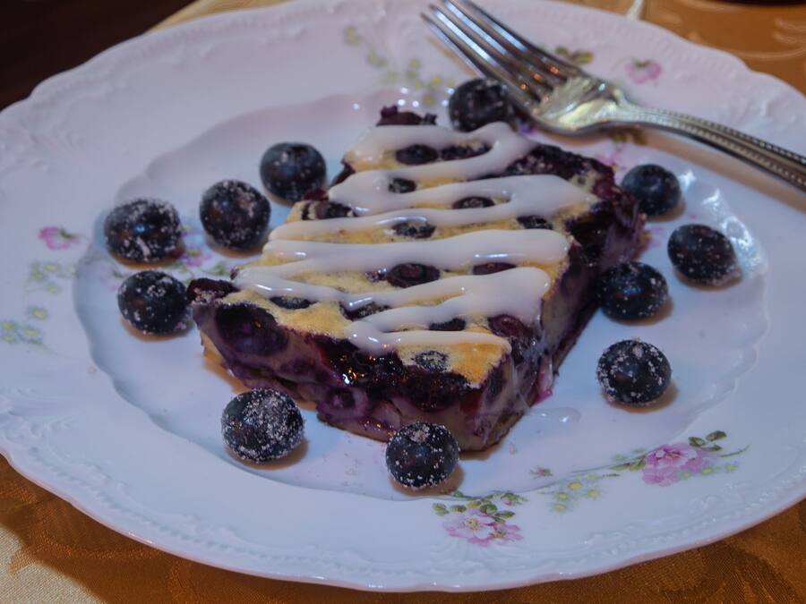 Una porción de tarta azul de moras sobre un plato con motivos florales, junto con moras, un tenedor, un vaso de agua y un jarrón decorativo al fondo.