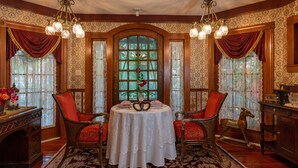 A dining room with a round table, two chairs, a chandelier, and a large stained glass window. - Cedar House Inn (St. Augustine)