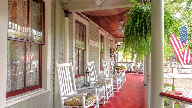 A covered porch with white chairs and red flooring, featuring American flags and hanging plants. - Cedar House Inn (St. Augustine)