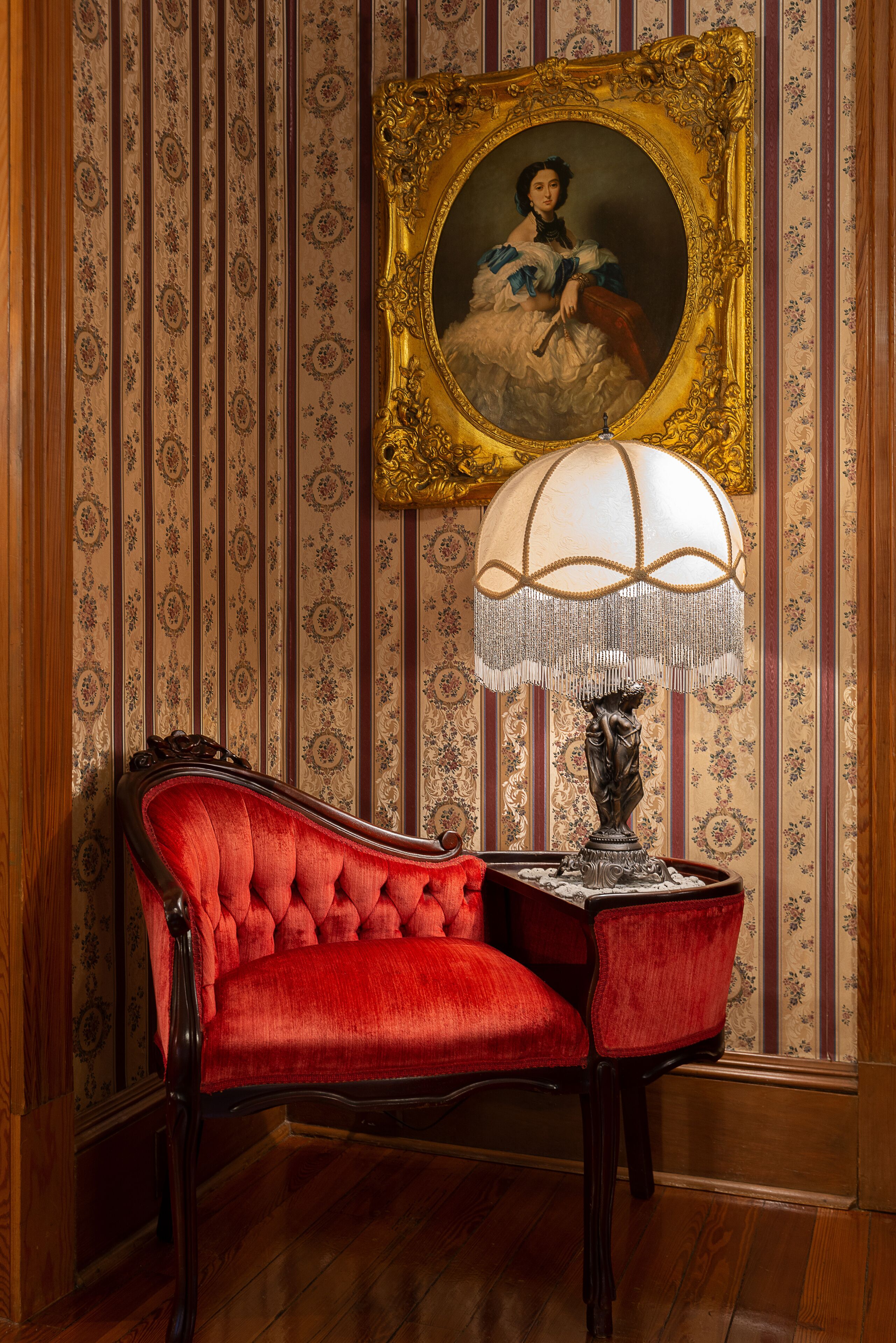 Ein Zimmer mit einem roten, gepolsterten Sessel, einem Tisch mit einer Lampe und einem gerahmten Porträt an der Wand.