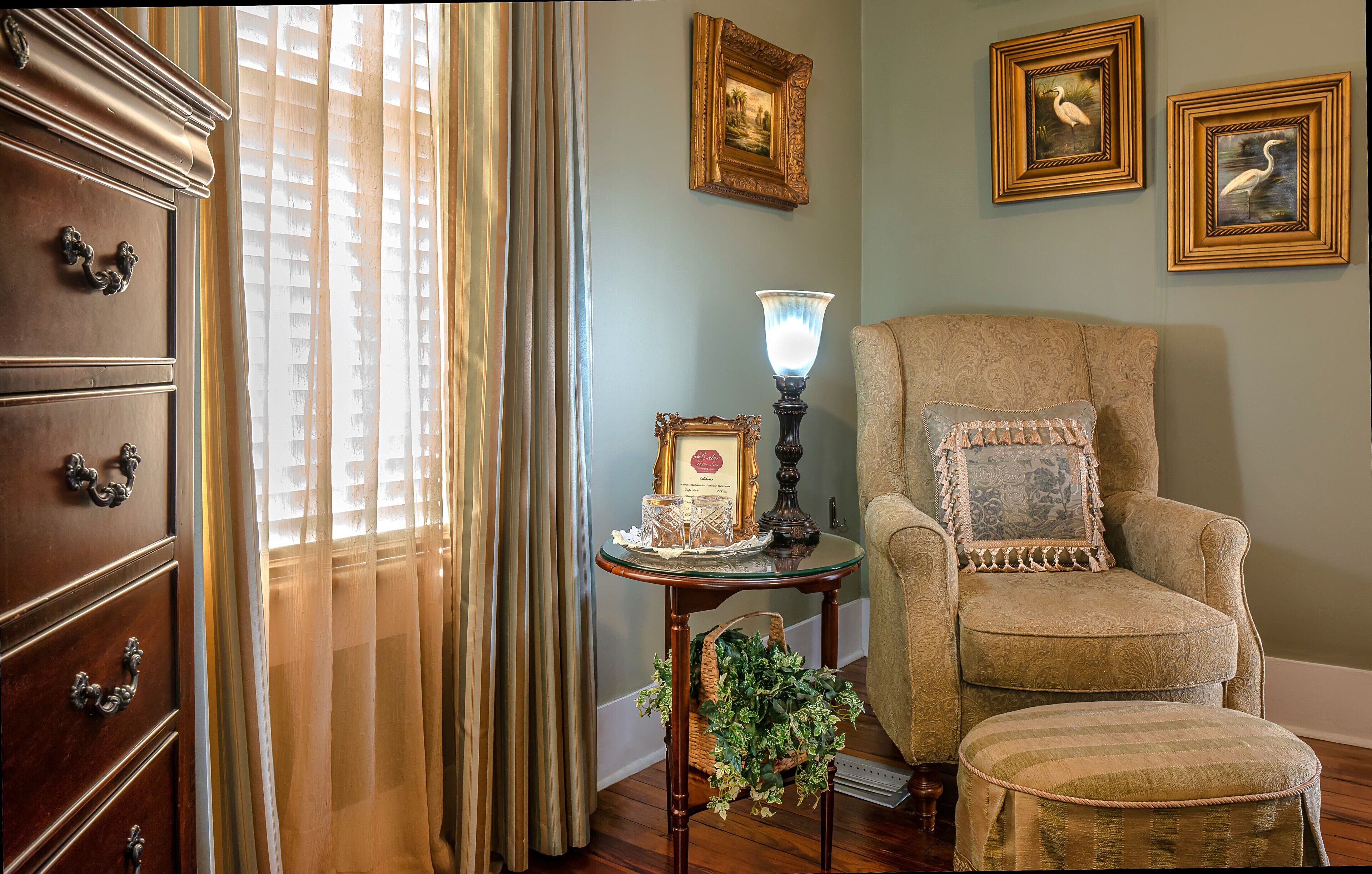 A room with a wooden dresser, a patterned armchair, a table with a lamp, and framed pictures on the wall.
