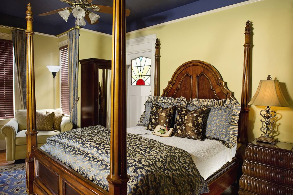 A four-poster bed with a canopy, a wooden nightstand with a lamp, and a view of a door with a stained glass window.