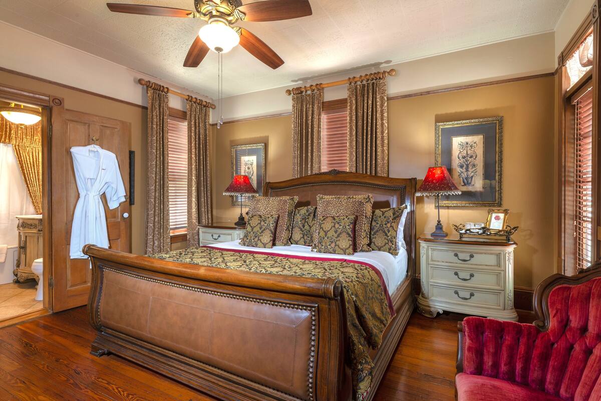 A bedroom with a wooden bed, two bedside tables, a red chair, a ceiling fan, and a window with curtains.