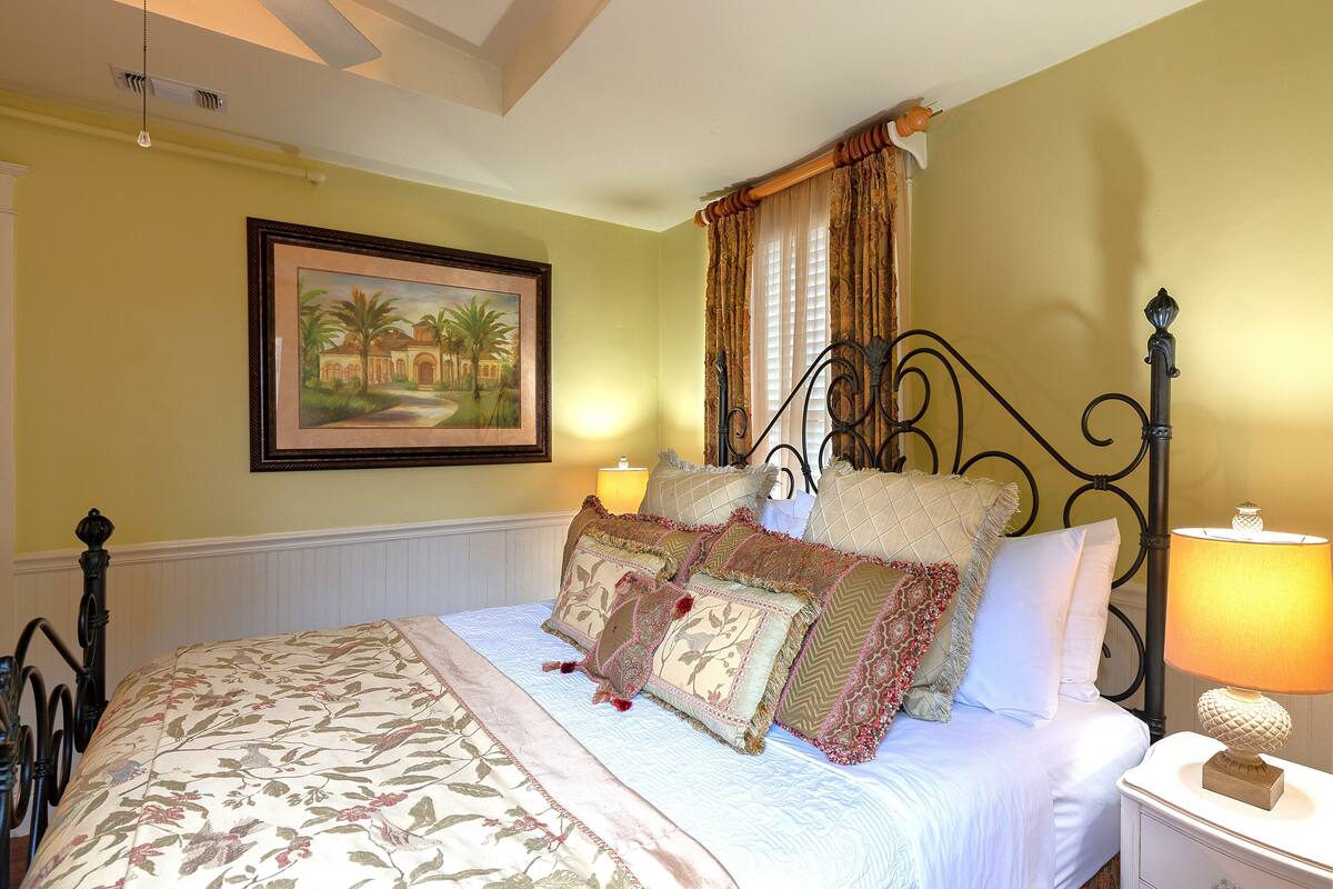 A bedroom with a bed, pillows, a nightstand, a lamp, a painting, and a window with curtains.