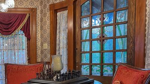 A room with a chess table, two chairs, a large window with stained glass, and patterned wallpaper. - Cedar House Inn (St. Augustine)