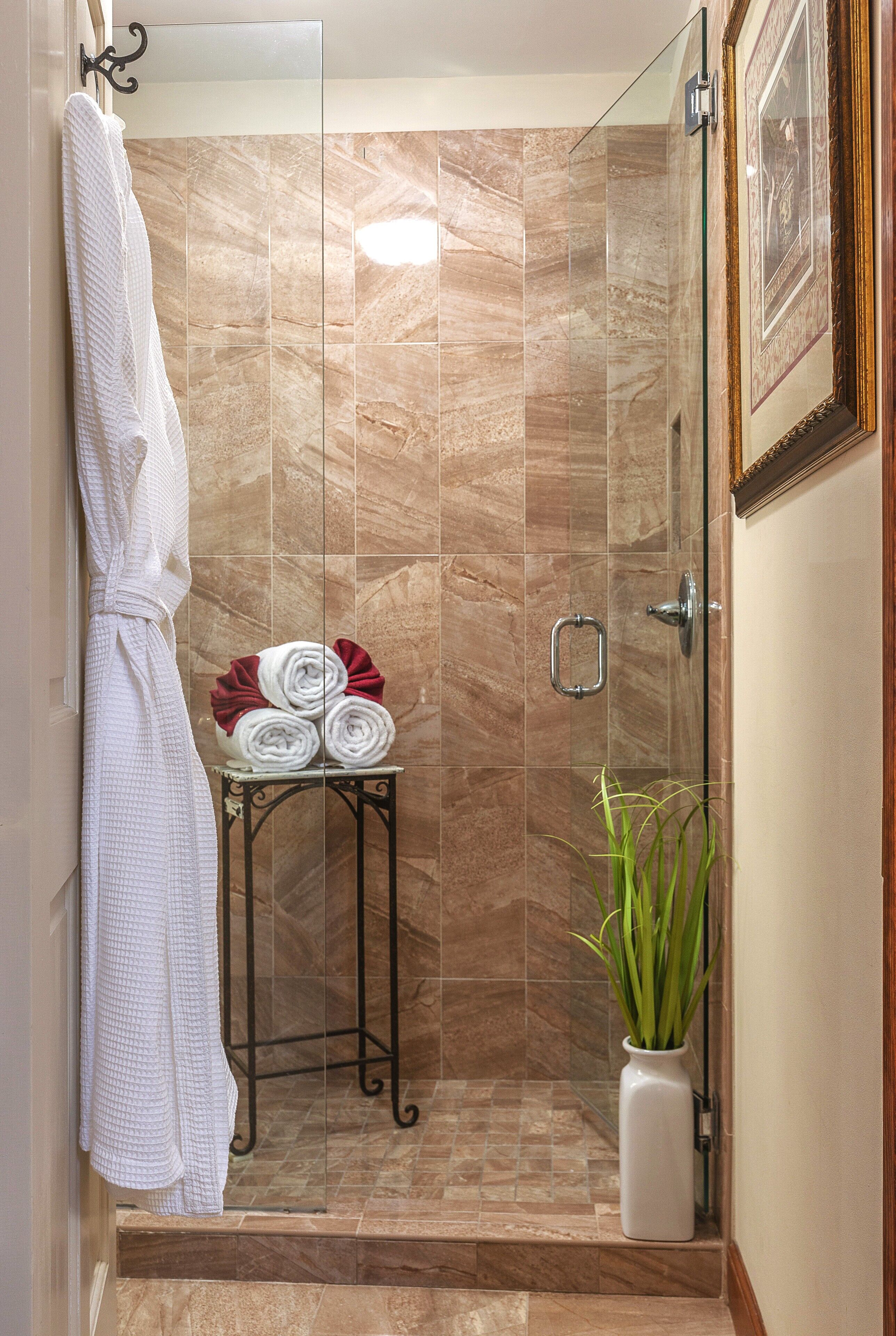 A glass-enclosed shower with marble tiles, a white bathrobe, a small table with rolled towels, and a potted plant.