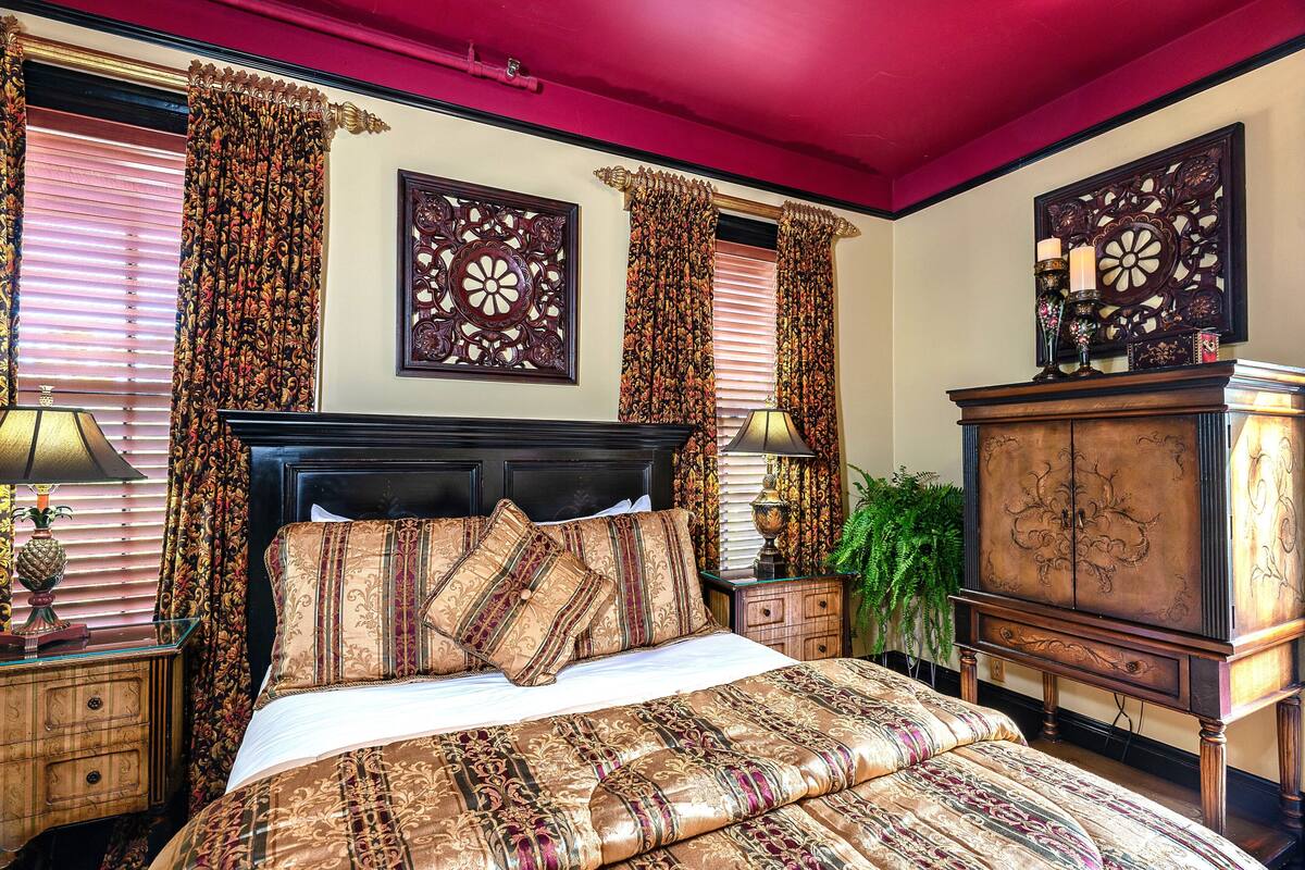 A bedroom with a large bed, ornate curtains, a wooden headboard, and a nightstand with a lamp.