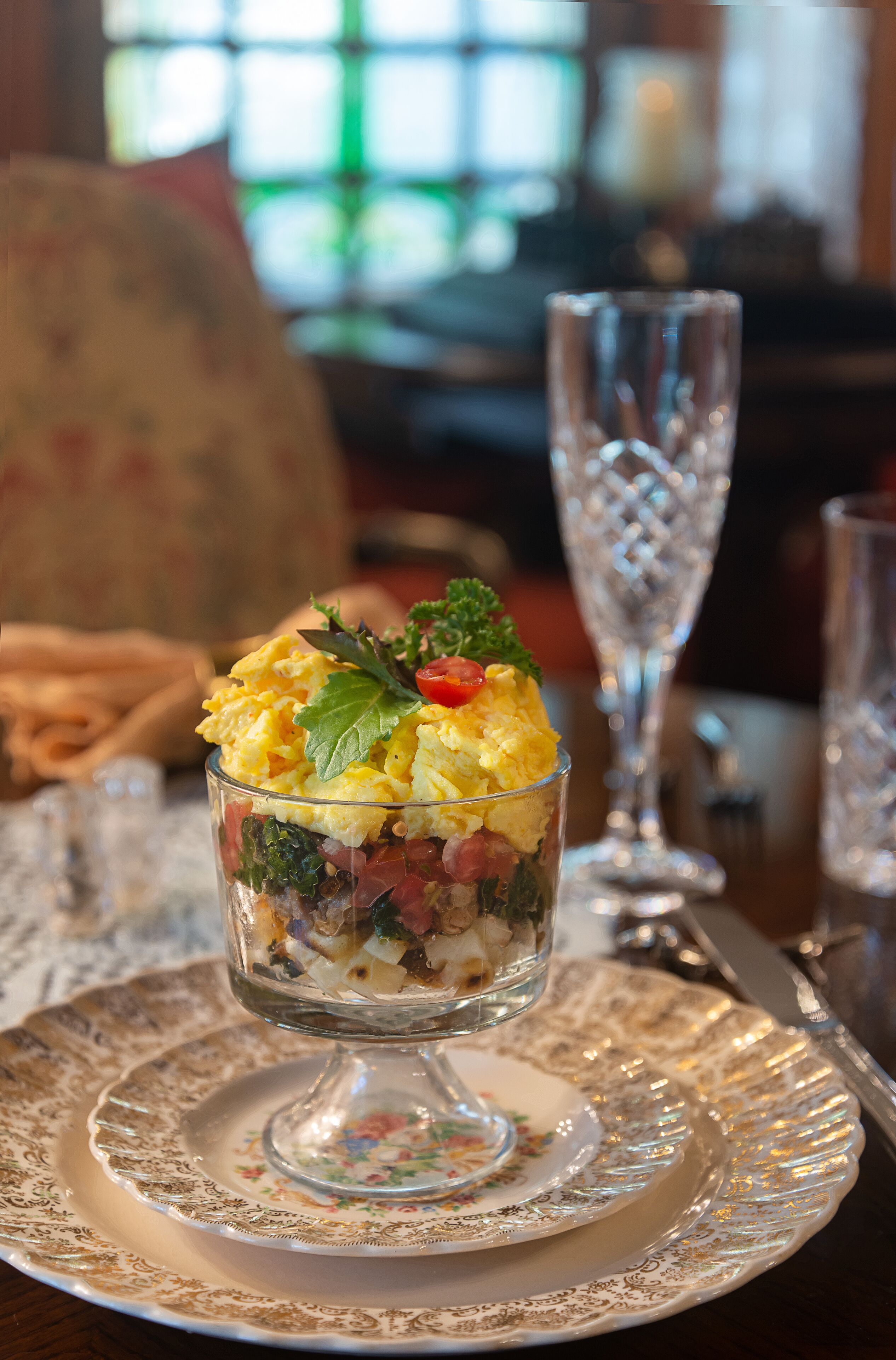 Postre en un cuenco de vidrio sobre un plato con estampado floral, acompañado de un vaso de agua y un tenedor sobre una mesa.