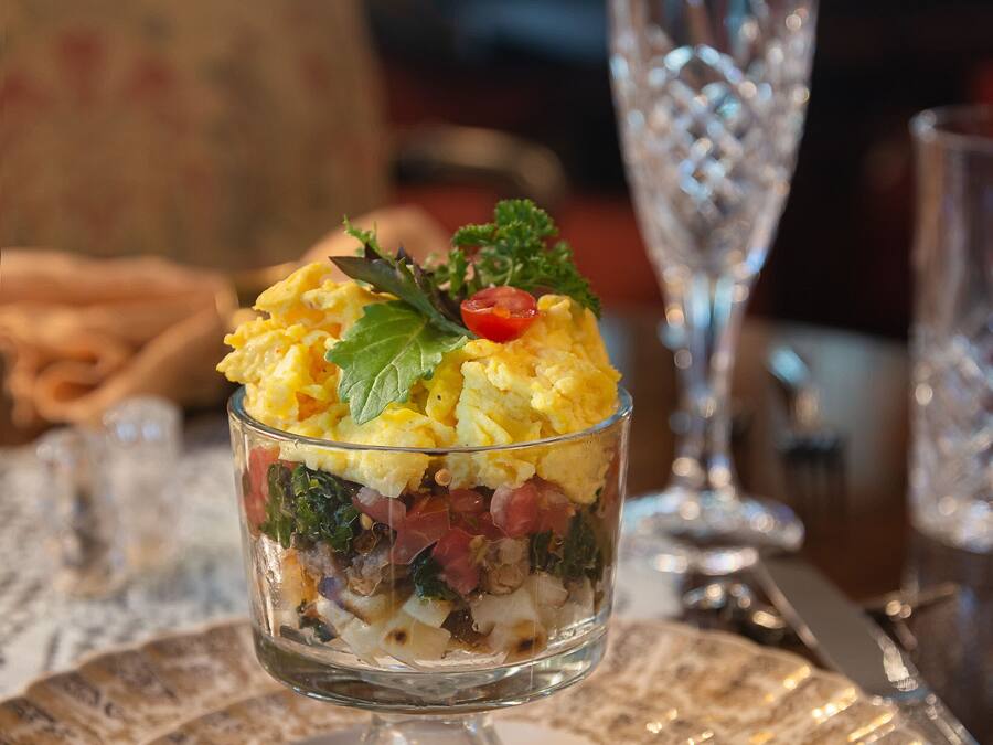 Postre en un cuenco de vidrio sobre un plato con estampado floral, acompañado de un vaso de agua y un tenedor sobre una mesa.