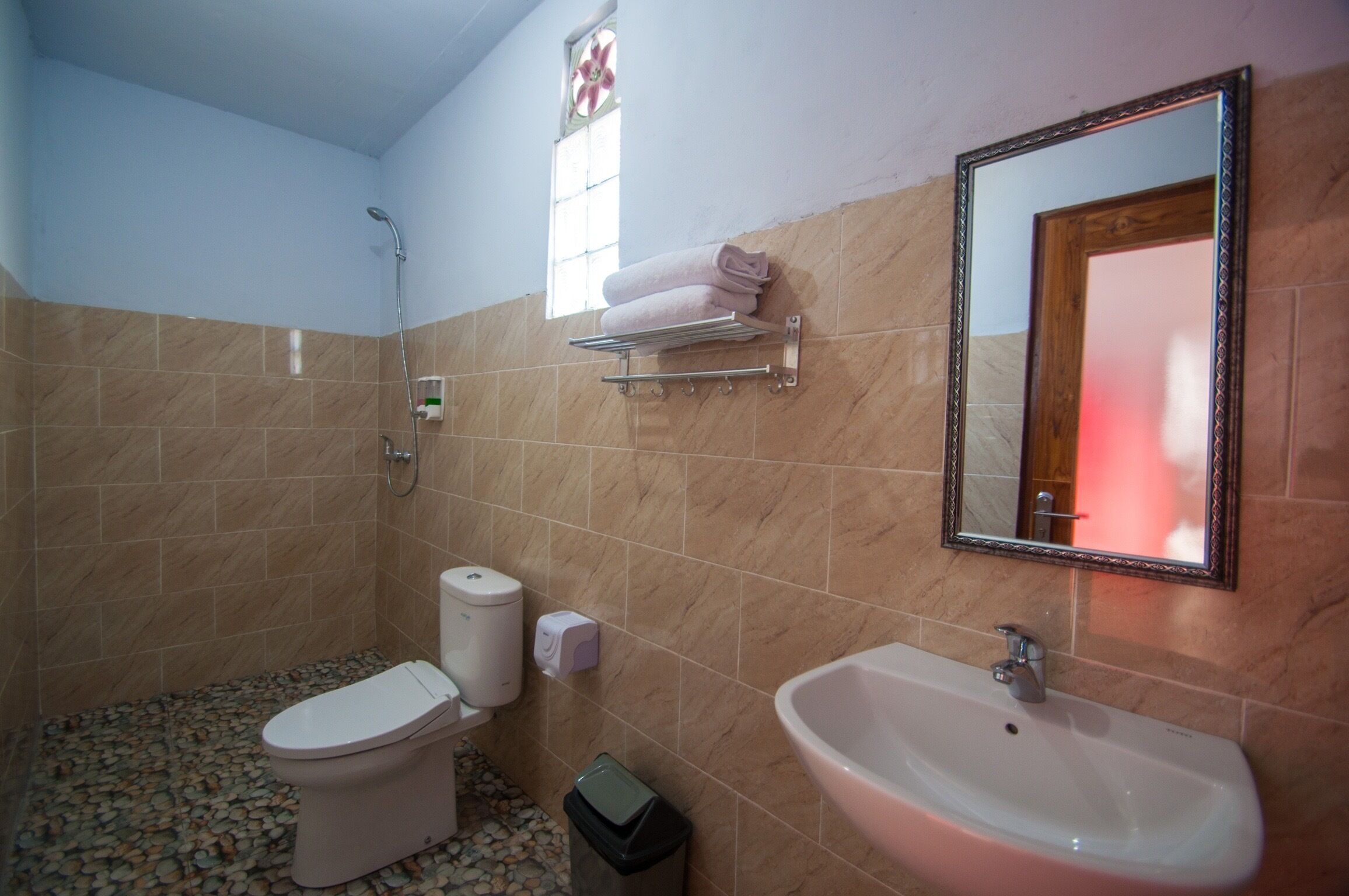 A bathroom with a toilet, sink, and shower. There is a towel rack with towels, a soap dispenser, and a trash bin.