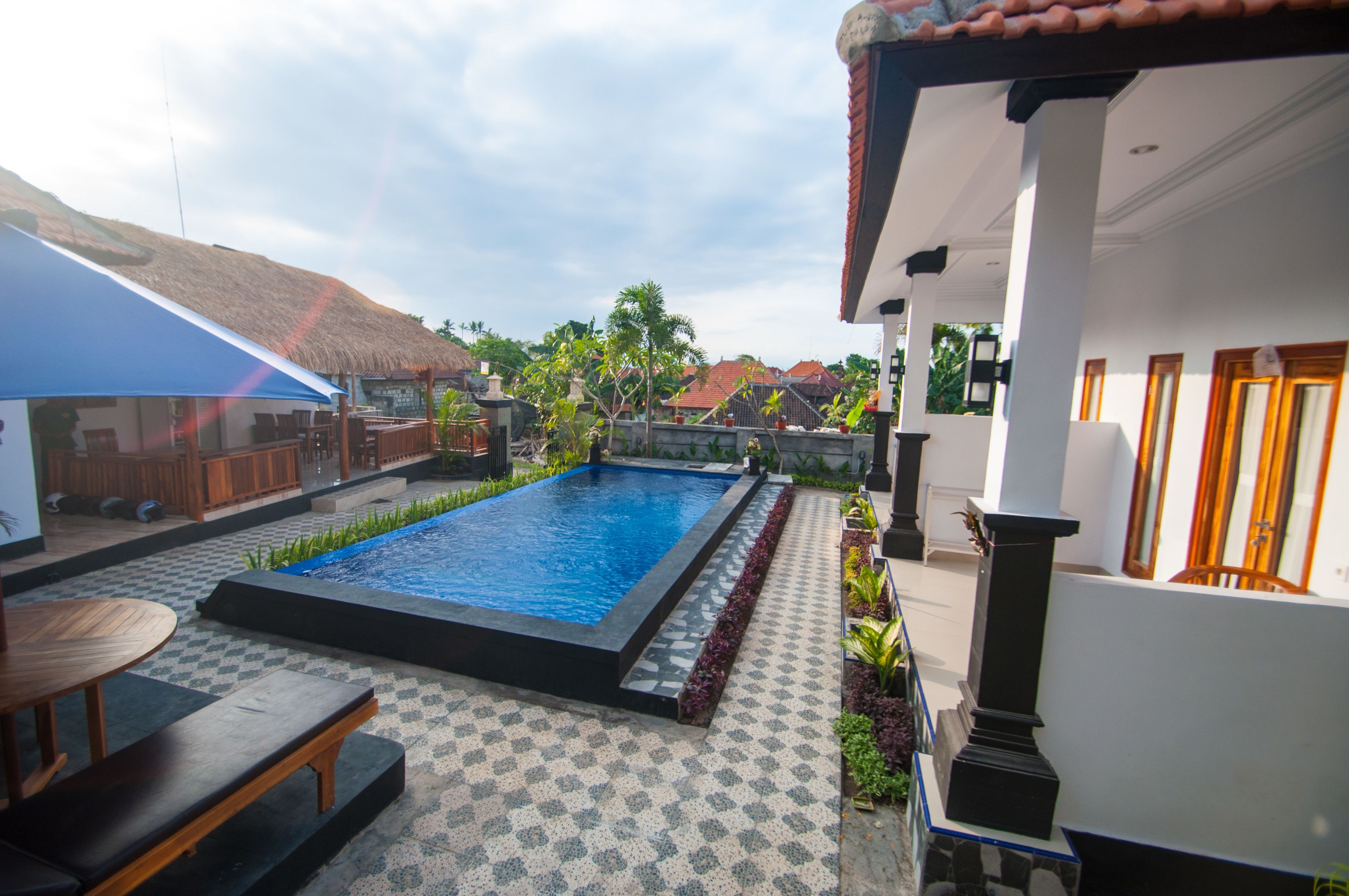 A swimming pool with a tiled deck, a wooden bench, and a thatched-roof structure in the background.
