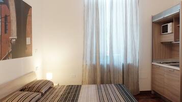 Un dormitorio con cama, ventana con cortinas translúcidas, área de cocina con microondas y un cuadro en la pared.