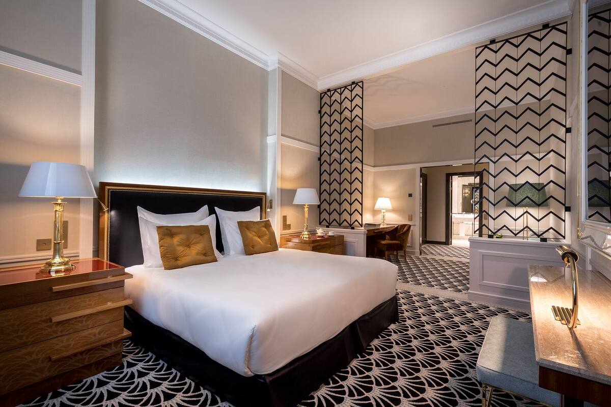 A hotel room with a large bed, two bedside tables with lamps, a desk with a chair, and a patterned carpet.