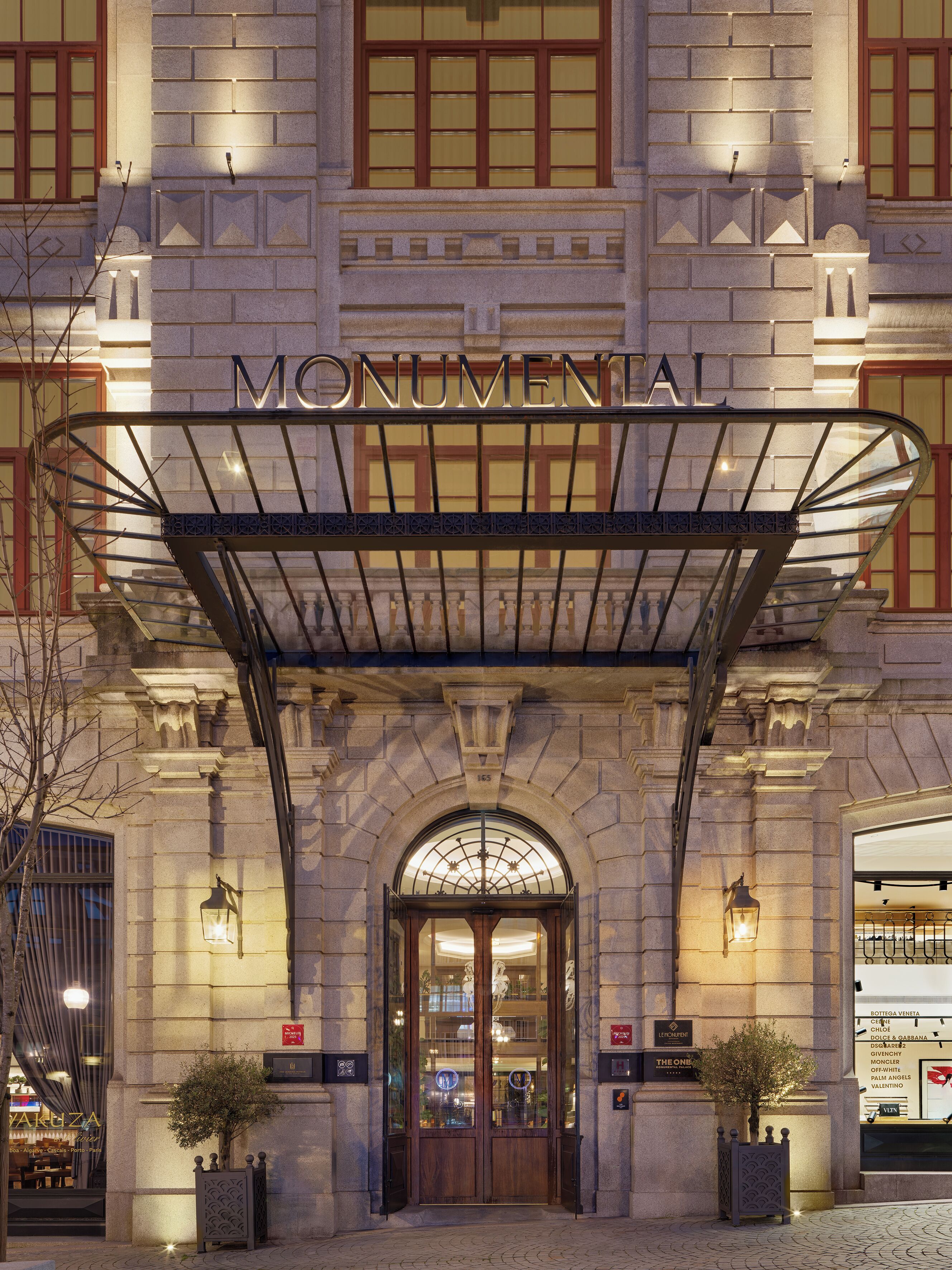 Entrada de hotel con un vestíbulo techado, un letrero que dice 'MONUMENTAL' y vista de la calle con un árbol y un anuncio de 'Bottega Veneta'.