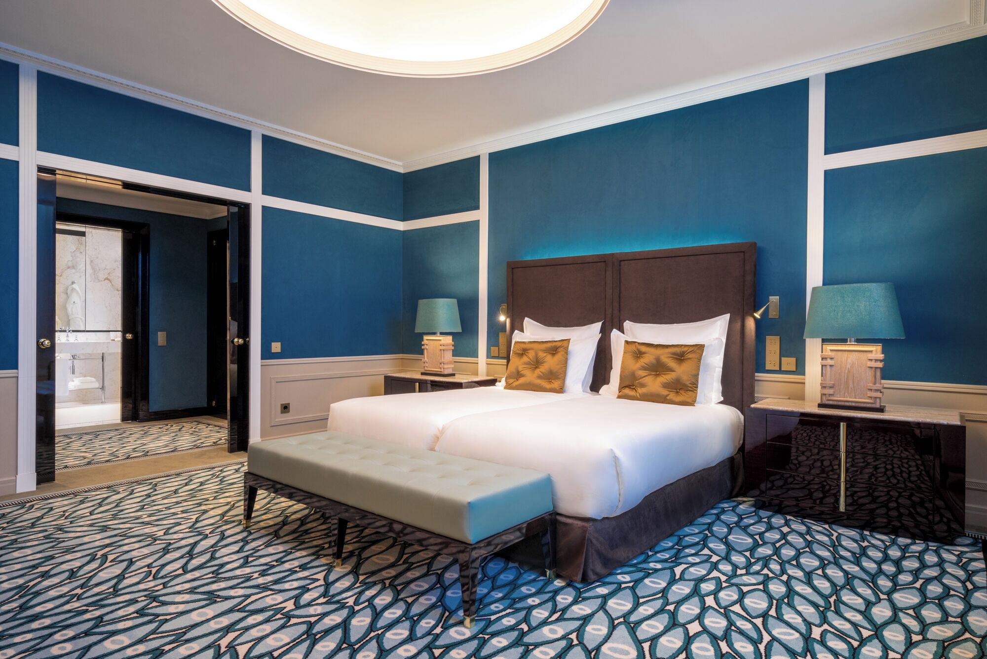 Habitación de hotel con una cama grande, un banco, dos mesitas de noche con lámparas y un baño visible a través de una puerta abierta.