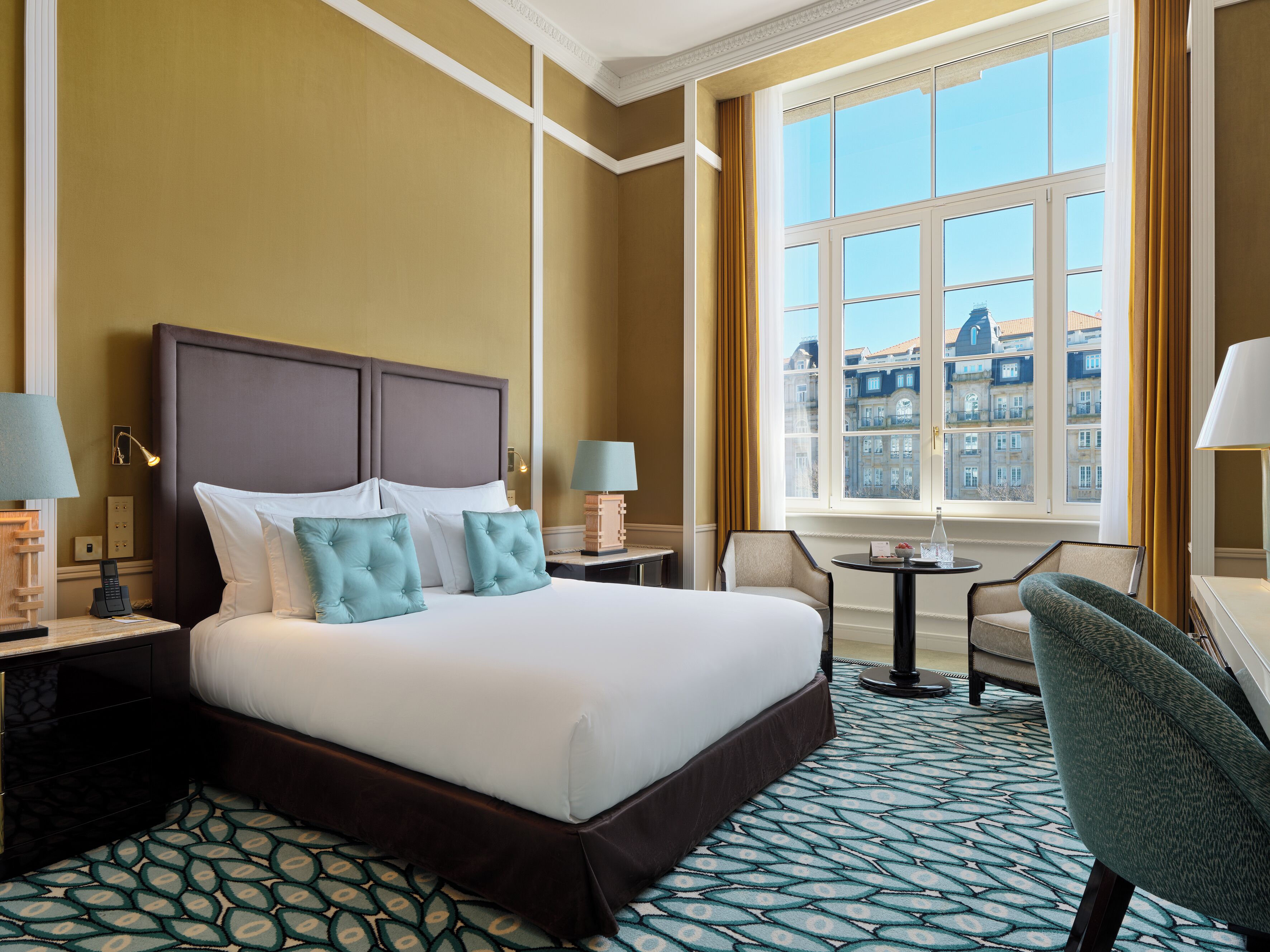Habitación de hotel con una cama grande, dos mesitas de noche, una silla, una pequeña mesa y un ventanal con vistas a edificios.