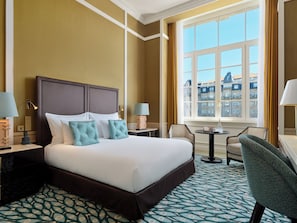Une chambre d’hôtel avec un grand lit, deux tables de chevet, une chaise, une petite table et une fenêtre donnant sur des immeubles.