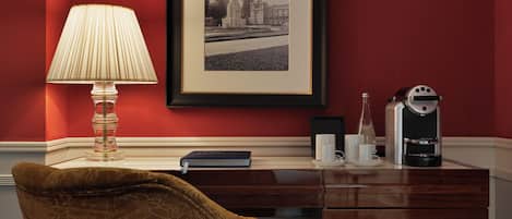 A room with a red wall, a framed black and white photo, a desk with a lamp, a chair, and a coffee machine.