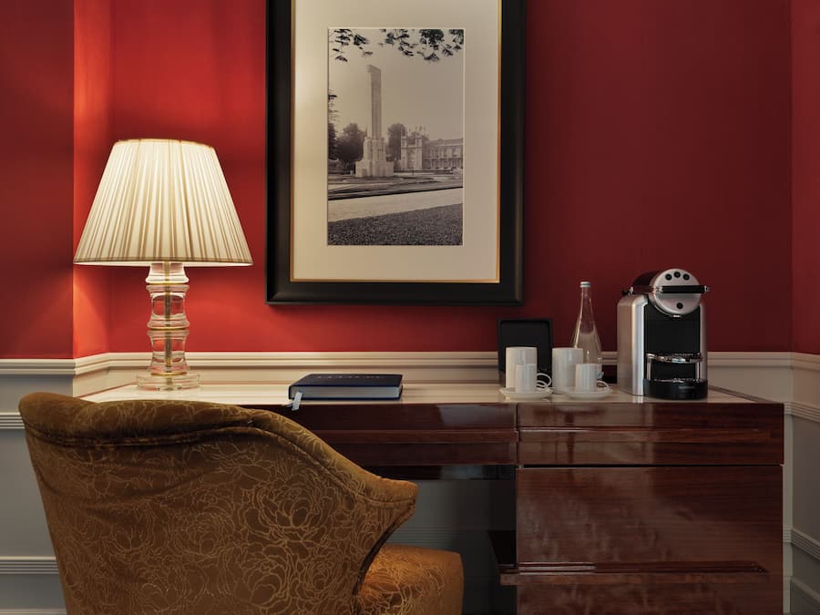 Una habitación con una pared roja, una fotografía blanco y negro enmarcada, un escritorio con una lámpara, una silla y una cafetera.