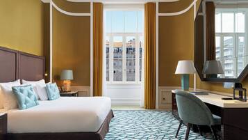 Habitación de hotel con cama, escritorio, silla, ventana con cortinas y vistas a edificios.