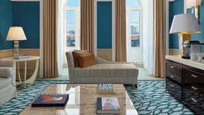 Habitación de hotel con un ventanal grande, un sofá, una mesita de centro con libros, una lámpara y un minibar.
