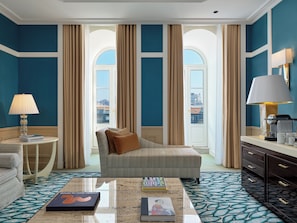 Habitación de hotel con un ventanal grande, un sofá, una mesita de centro con libros, una lámpara y un minibar.