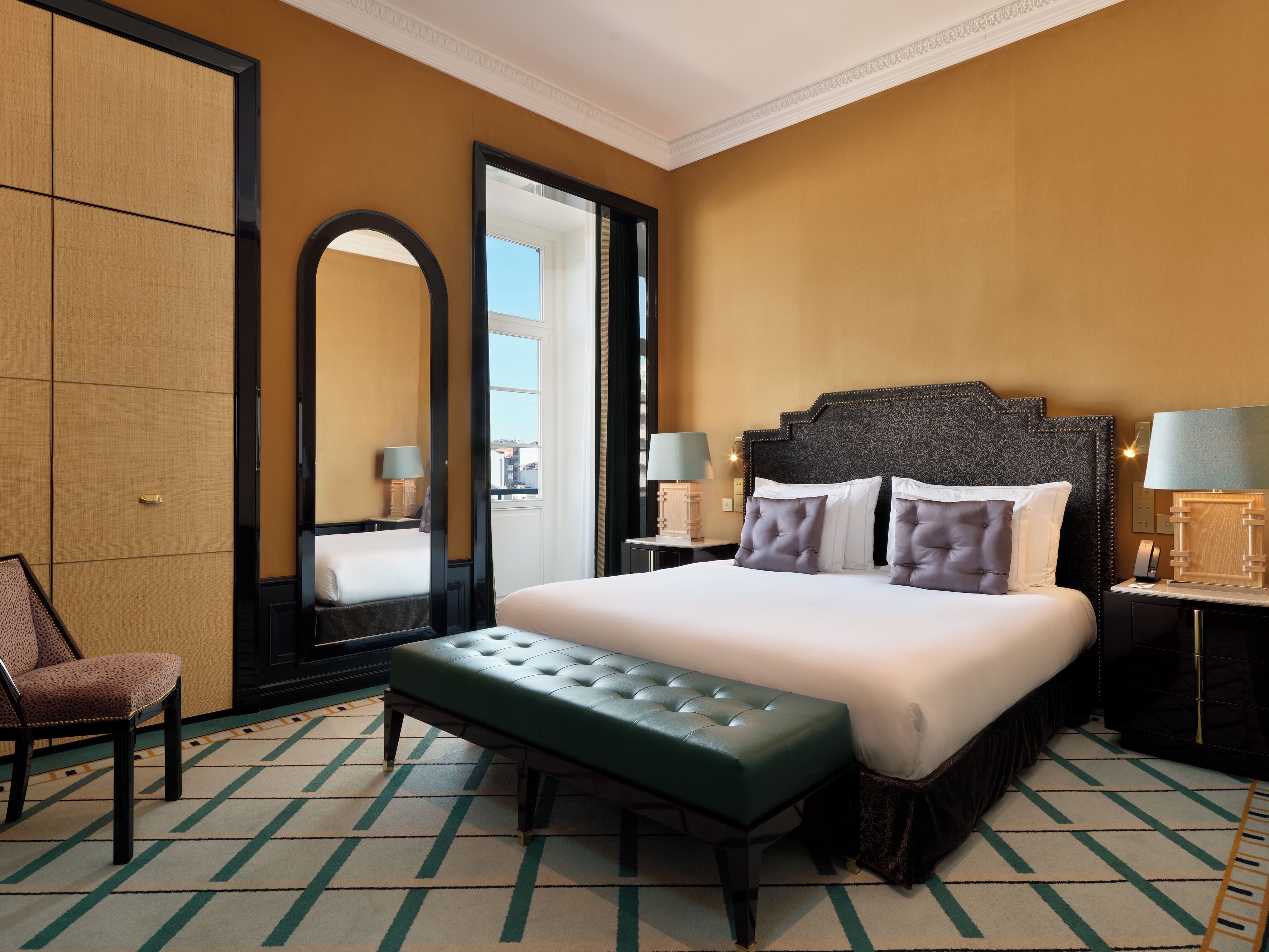 Habitación de hotel con una cama grande, un banco, una silla, un espejo y dos mesitas de noche con lámparas.