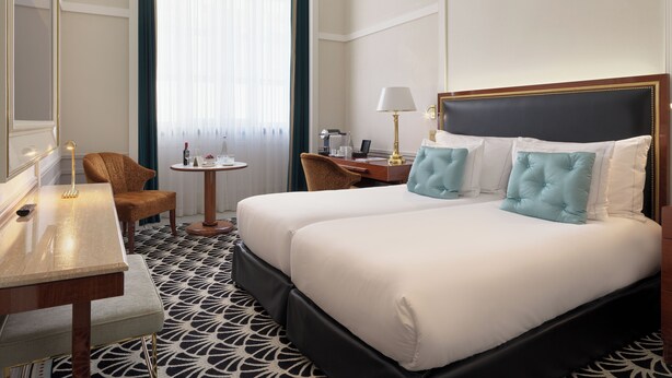 Habitación de hotel con cama, mesitas de noche, un escritorio, una silla, una pequeña mesa con lámpara y una ventana con cortinas.