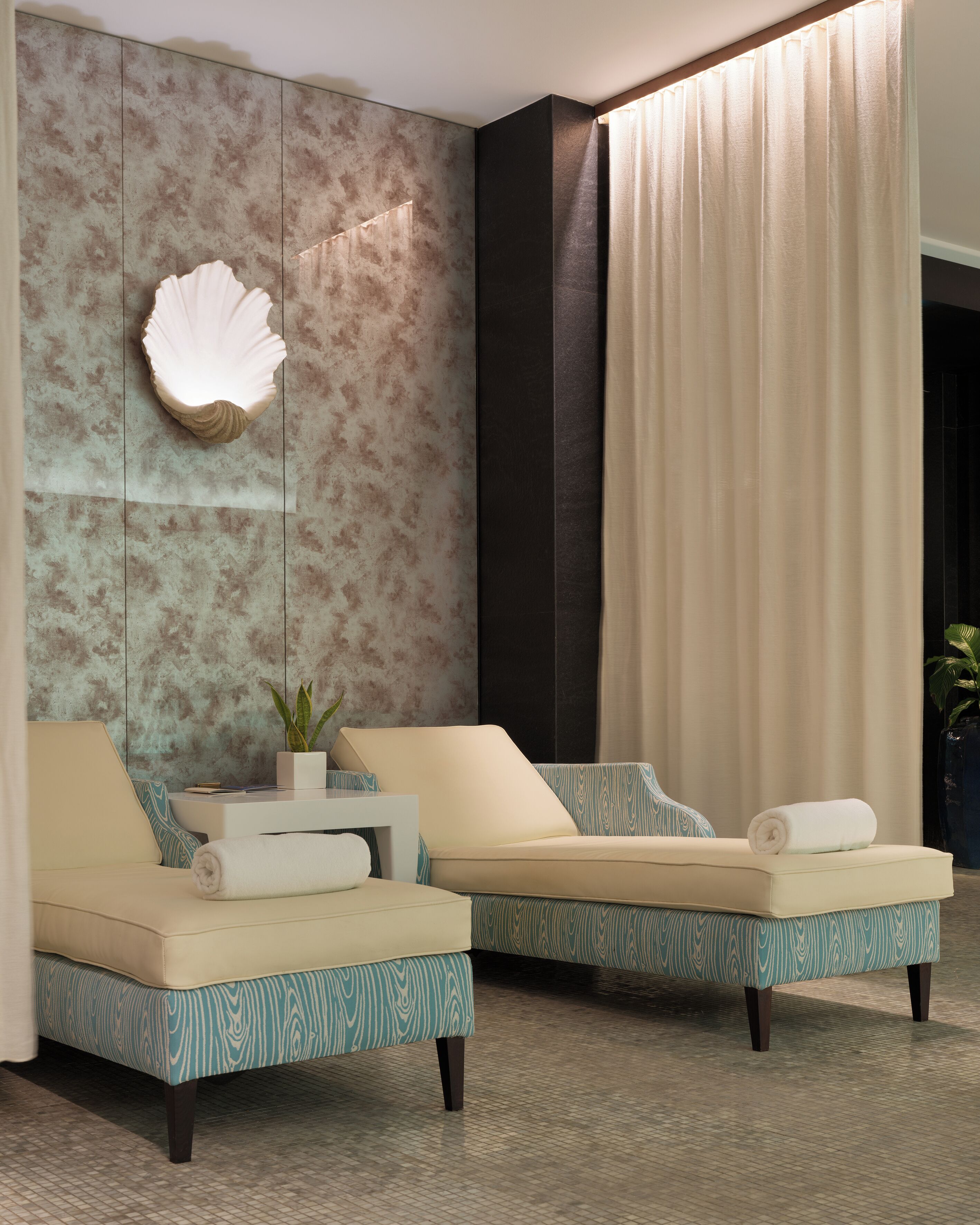 Quarto de hotel moderno com duas sofás, um detalhe decorativo de conchas na parede, um vaso de plantas e uma grande janela com cortinas.